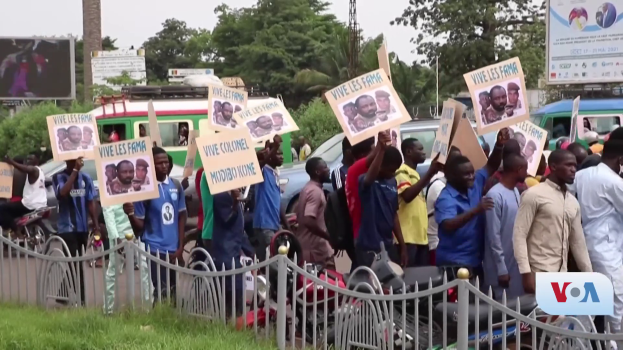 Eine Gruppe von Menschen mit Schildern steht vor einem Zaun, umgeben von Fahrzeugen, Bäumen, Polen, Plakaten und einem Bildschirm, mit Gras am Boden und dem Himmel im Hintergrund, scheinbar protestierend gegen eine Regierungsentscheidung.
