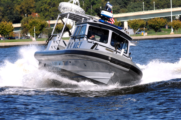 Ein Polizeiboot fährt über ein Gewässer mit einer Person darin, mit einer Brücke, Bäumen, Polen, Fahrzeugen, Gras und einem klaren blauen Himmel im Hintergrund.