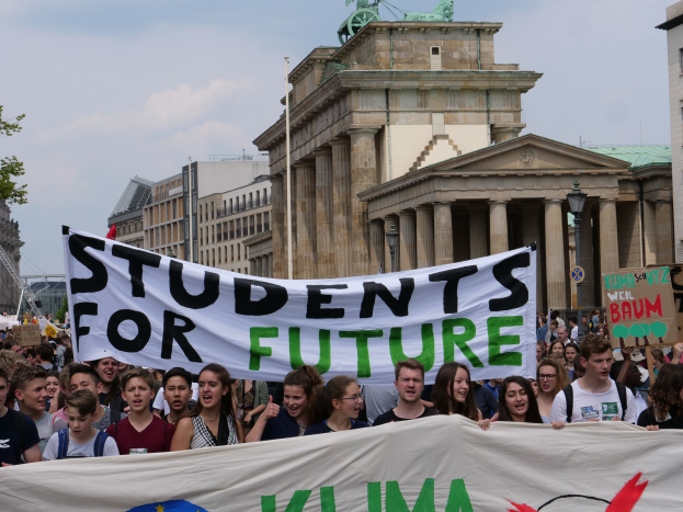 Eine Gruppe von Studenten marschiert in Berlin, die eine bunt bemalte Schrifttafel tragen, auf der "Students for Future" steht, mit Gebäuden, Bäumen und Himmel im Hintergrund.