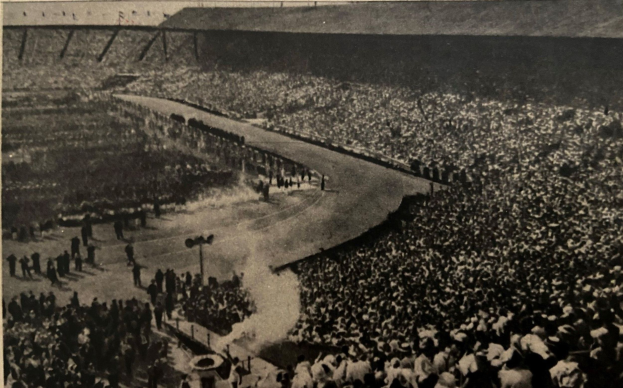 Ein altes Schwarz-Weiß-Foto einer großen Menge in einem Stadion, mit einer zentralen Straße, die von Pfählen gesäumt ist, und Text am unteren Rand.