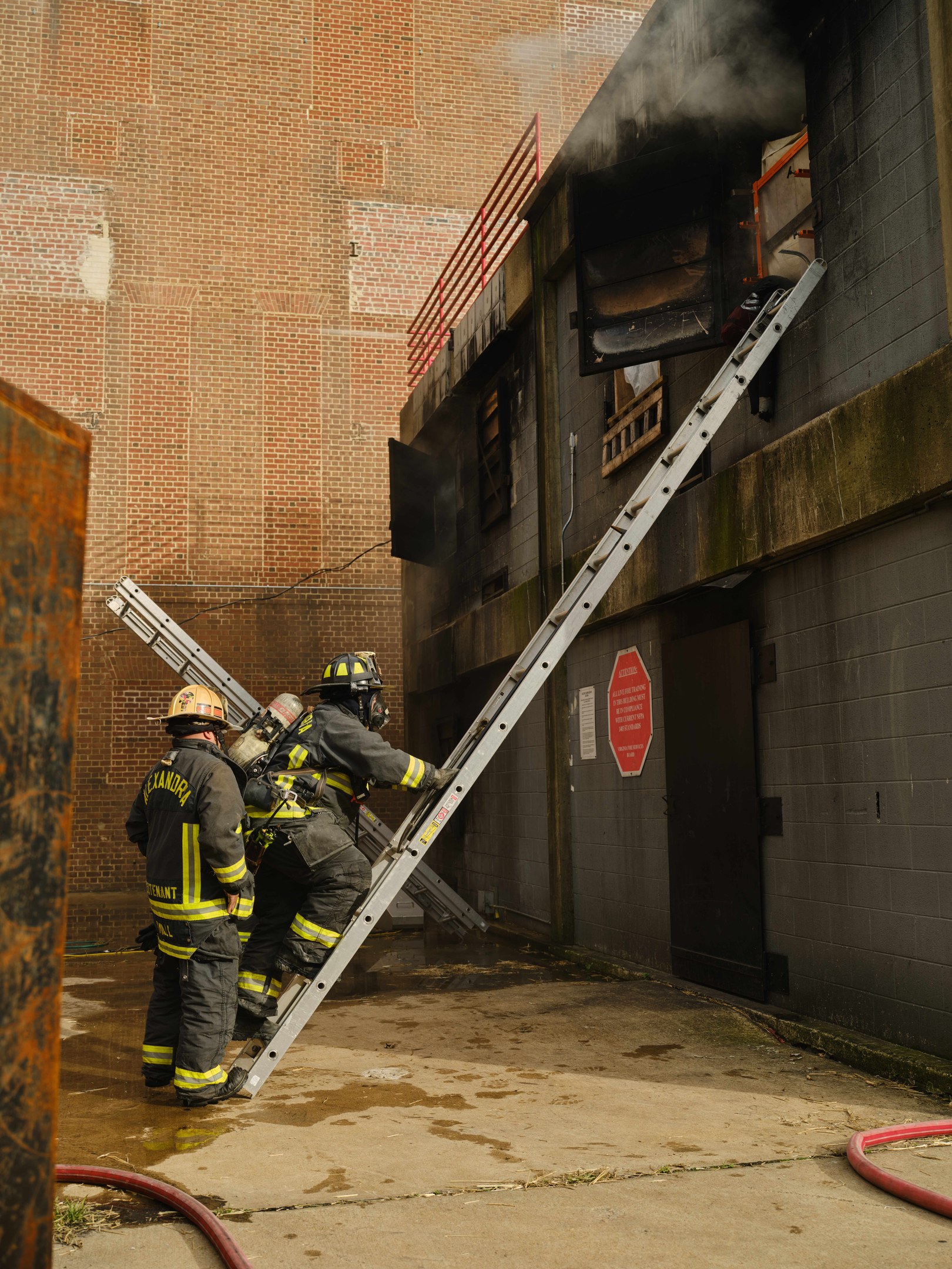 Feuerwehrleute mit Helmen klettern an einer Leiter zu einem raucherfüllten Gebäude hoch, mit einem Metallobjekt links und Rohren auf dem Boden.