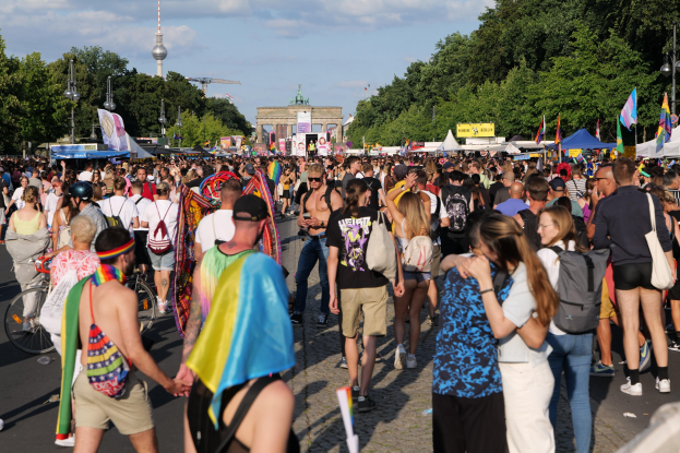 Eine große Menschenmenge marschiert durch eine Straße in Berlin, Deutschland, mit vielen, die Mützen tragen, Taschen tragen und Fahnen halten, neben Zelten, Bannern, Laternenmasten, Bäumen, Gebäuden und einem Turm unter einem bewölkten Himmel.