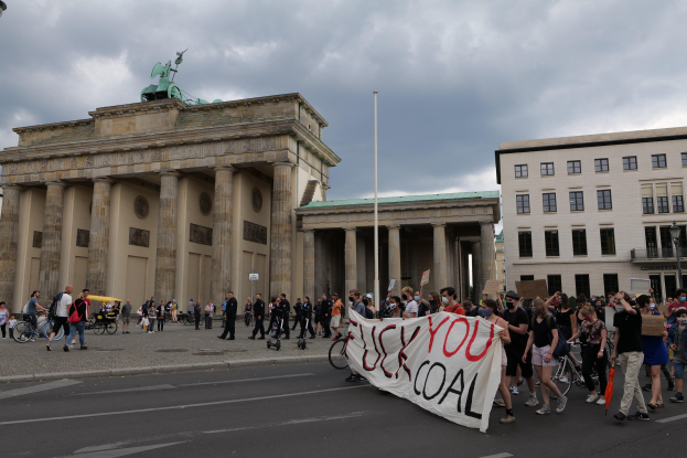 Eine Gruppe von Menschen zu Fuß und mit dem Fahrrad auf einer Straße vor dem Brandenburger Tor in Berlin, die eine Fahne halten, mit sichtbaren Säulen und Fenstern des Tors im Hintergrund.