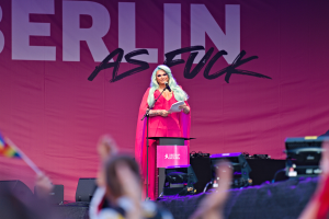 Eine Frau in einem pinken Kleid steht an einem Podium und hält ein Mikrofon und Papier in den Händen, während sie zu einer Menge spricht, die ein 'Berlin as Fuck'-Plakat im Hintergrund hat.