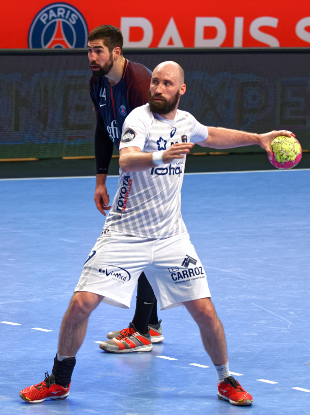 Zwei Männer beim Handballspielen auf einem Platz, einer hält den Ball, mit einer Tafel im Hintergrund, auf der "Paris Saint-Germain vs Paris Saint Germain" steht.