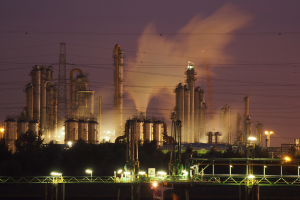 Ölraffinerie bei Nacht mit Rauch, beleuchtet von benachbarten Lichtern, mit sichtbarem Himmel und einem Wasserzeichen.