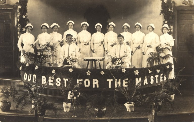 Eine Gruppe von Frauen in weißen Kleidern und Hauben steht auf einer Bühne mit einem Banner, auf dem 'Unser Bestes für den Meister' steht, umgeben von Blumentöpfen, Türen und einer Wand.