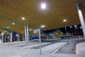 Bus-Haltestelle bei Nacht mit Deckenbeleuchtung, Fahrzeuge auf der Straße, Beschilderung, einige Menschen und beleuchtete Gebäude im Hintergrund.