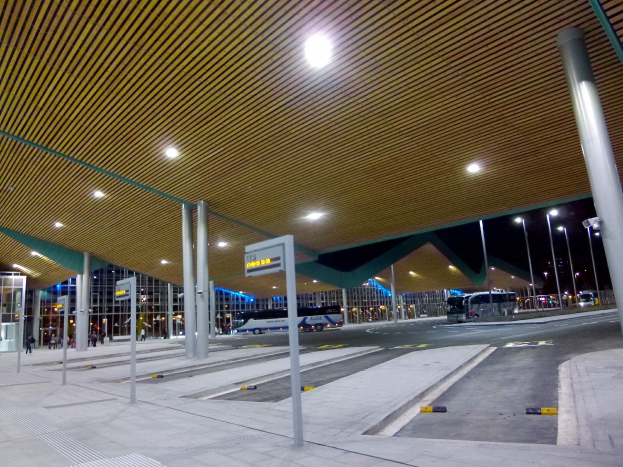 Bus-Haltestelle bei Nacht mit Deckenbeleuchtung, Fahrzeuge auf der Straße, Beschilderung, einige Menschen und beleuchtete Gebäude im Hintergrund.