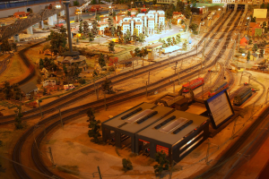 Eine Modellbahnanlage in einem Museum mit Gebäuden, Bäumen, Eisenbahnschienen, Fahrzeugen, Polen, Lichtern, Platten und anderen Gegenständen, die eine belebte Stadtlandschaft schaffen.