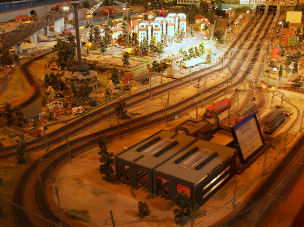 Eine Modellbahnanlage in einem Museum mit Gebäuden, Bäumen, Eisenbahnschienen, Fahrzeugen, Polen, Lichtern, Platten und anderen Gegenständen, die eine belebte Stadtlandschaft schaffen.