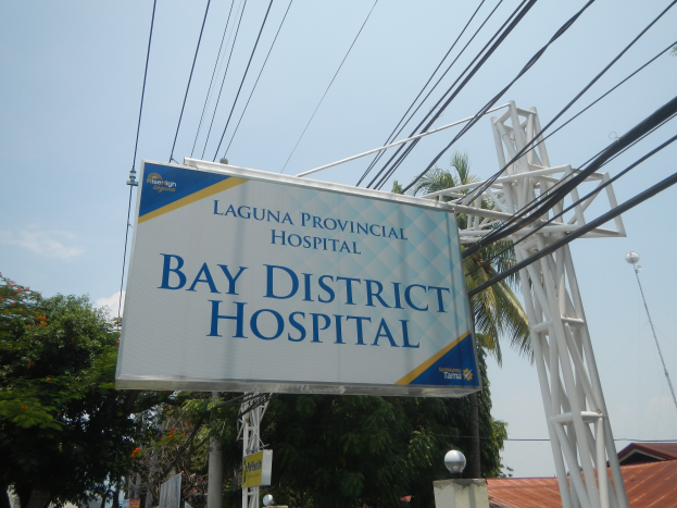 Krankenhausgebäude im Landkreis mit einem Schild, Strommasten, Laternen, Bäumen und anderen Gebäuden unter einem bewölkten Himmel.