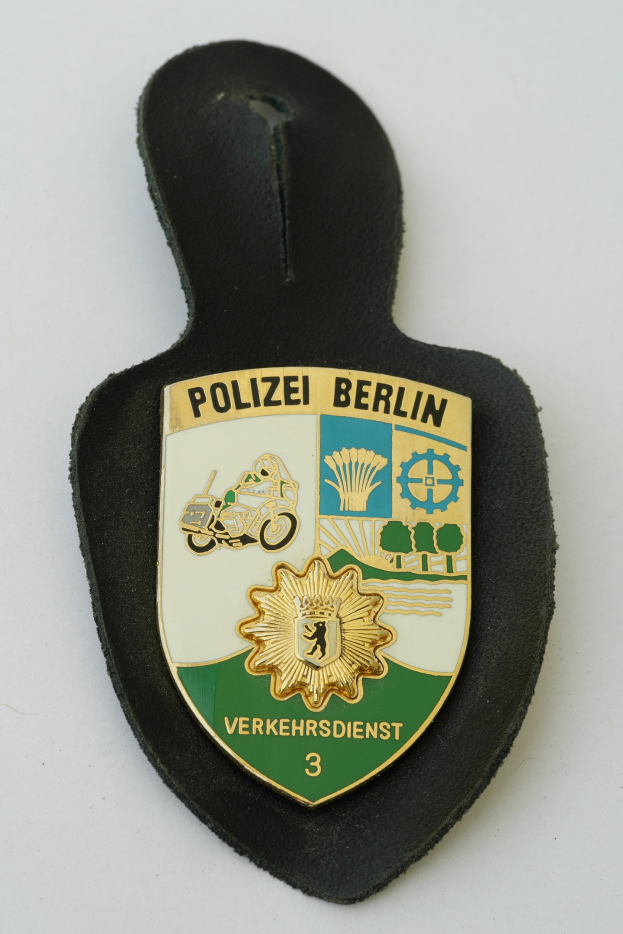 Ein schildförmiges Polizeibadge mit "Polizei Berlin" in schwarzer Schrift auf einem blauen Hintergrund mit einem weißen Stern und Rahmen, das auf einer Oberfläche liegt.