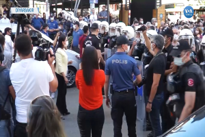 Eine Gruppe von Menschen steht vor einer Reihe von Polizisten mit Helmen und Kameras, mit Fahrzeugen auf der Straße und Gebäuden, Bäumen und Schildern mit der Aufschrift "türkische Polizei bei Protesten in Ankara" im Hintergrund.