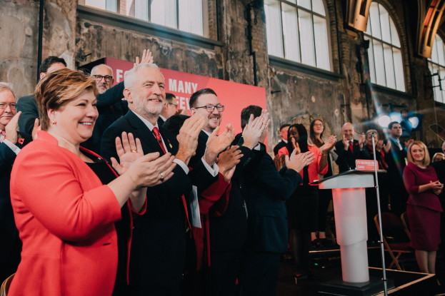 Eine Gruppe von Menschen, die vor einem Publikum stehen und jubeln, mit einem Podium, einem Mikrofon und einer Tafel auf der rechten Seite und Stühlen, einer Fahne, einer Wand, Fenstern und Lichtern im Hintergrund.