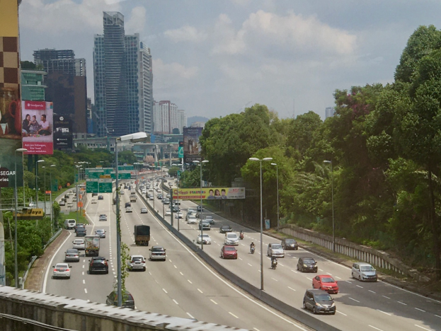Eine vielbefahrene Autobahn mit zahlreichen Fahrzeugen, gesäumt von hohen Geb├Ąuden, Straßenlaternen, Werbetafeln und B├Ąumen, unter einem bew├Âlktem Himmel.