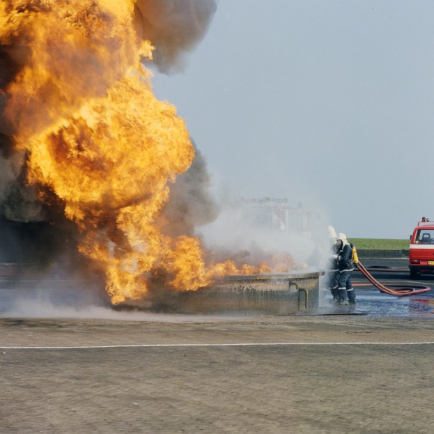 Ein Feuerwehrauto steht in Flammen am Straßenrand, zwei Personen mit Helmen und Schläuchen in der Nähe und ein Fahrzeug und der Himmel im Hintergrund.