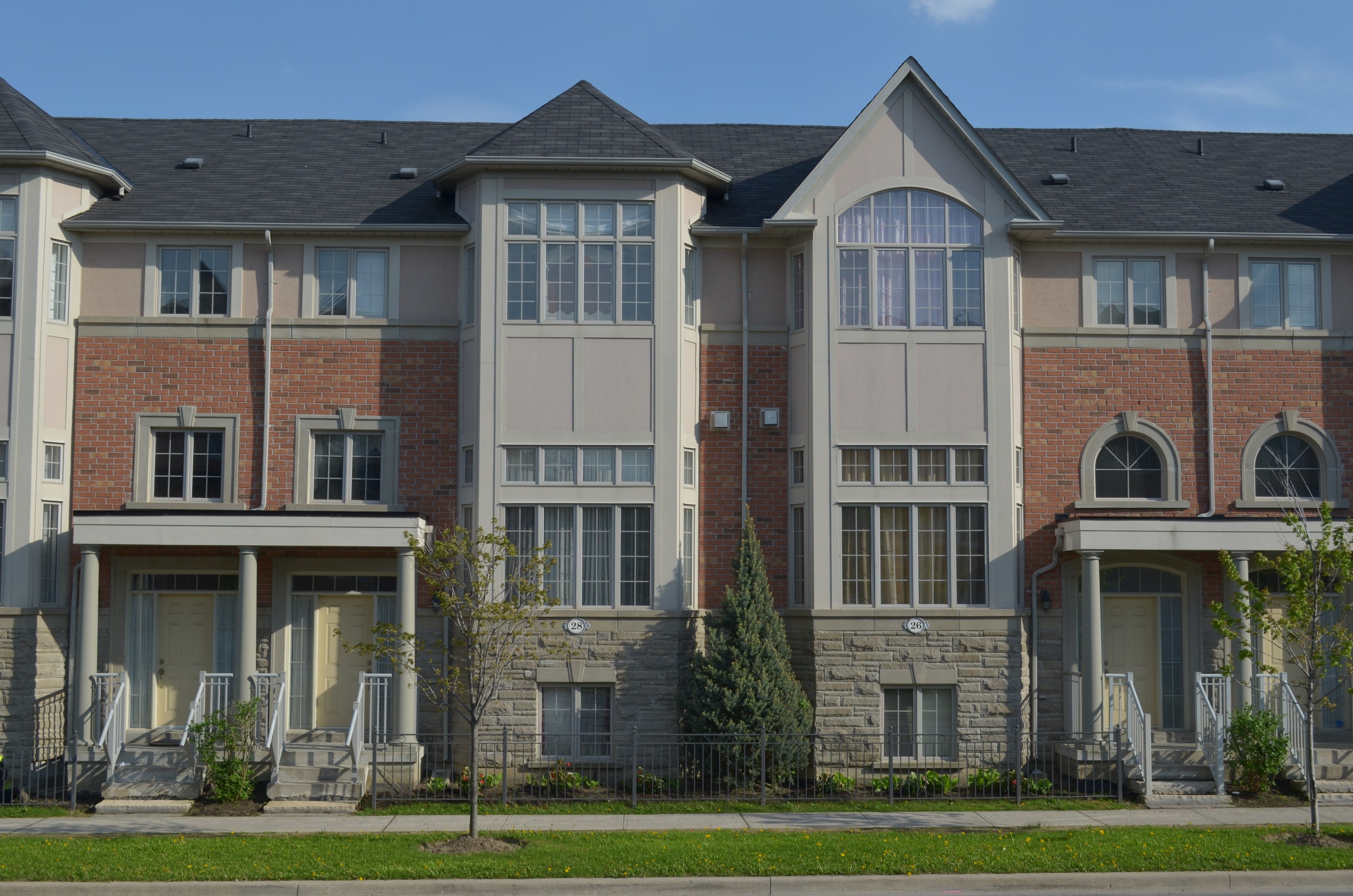 Eine Reihe von Stadthäusern in einer Wohngegend mit sichtbaren Fenstern, Türen, Säulen, Geländern, Stufen, Pflanzen, Bäumen, Gras, einer Straße im Vordergrund und dem Himmel im Hintergrund.