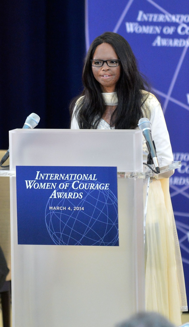 Eine Frau in einem weißen Kleid und Brille steht an einem Podium mit zwei Mikrofonen und spricht vor einer Menge, mit einem blauen Banner im Hintergrund, auf dem "Internationale Frauen des Mutes Awards 2014" steht.