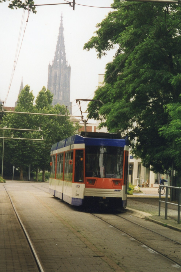 Rotes und weißes Zugfahrzeug auf Schienen neben einem hohen Gebäude, mit einer Person, die ein Fahrrad auf einem Fußweg neben den Schienen fährt, Bäume, die die Schienen säumen, und Gebäude im Hintergrund unter einem klaren blauen Himmel.