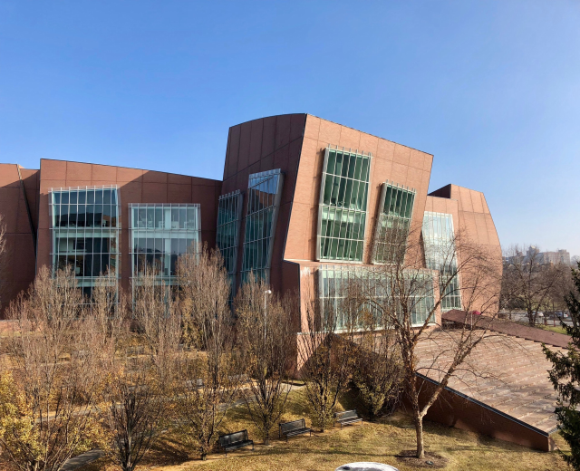 Großes modernes Gebäude mit vielen Fenstern, umgeben von Bäumen, Bänken, Gras und einem Laternenmast, mit anderen Gebäuden und einem klaren blauen Himmel im Hintergrund, identifiziert als der neue Campus der University of Illinois at Chicago.