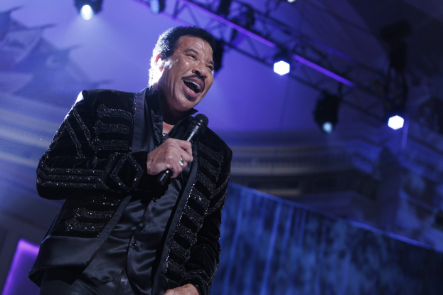 Lionel Richie performs bei den Grammys, trägt ein schwarzes Kleid, hält ein Mikrofon und hat eine dramatische Bühnenbeleuchtung.
