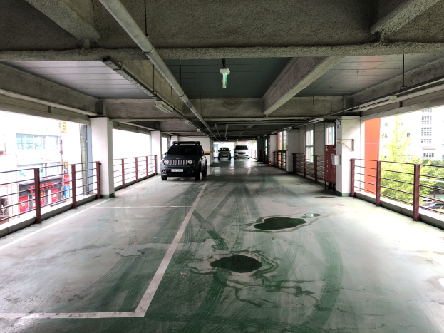 Ein mehrgeschossiges Parkhaus mit geparkten Autos, Gel├Ąndern, Pfeilern, Lichtern, Rohren und B├Ąumen, vor einer Stadtkulisse mit Geb├Ąuden mit Fenstern.