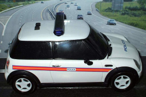 Ein weißer Polizeiwagen steht im Vordergrund mit mehreren Fahrzeugen, die sich dahinter auf der Straße bewegen, umgeben von Gras und Bäumen auf beiden Seiten, in einer ländlichen Umgebung.