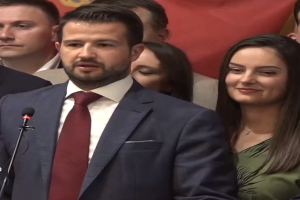 Ein Mann im Anzug spricht in ein Mikrofon an einem Rednerpult, mit einem Banner hinter ihm und einer Menge vor ihm.