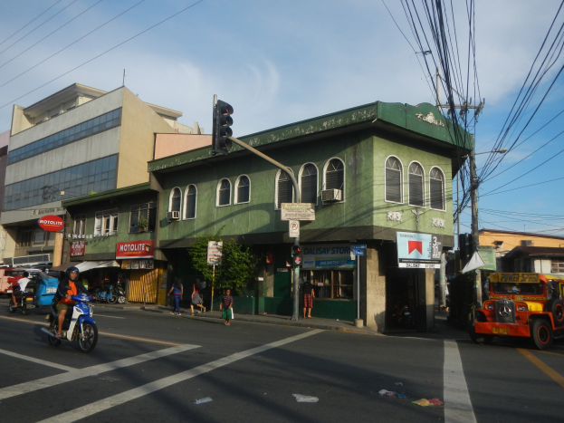 Eine belebte Stadtstraße mit einem grünen Gebäude im Hintergrund, Fahrzeugen auf der Straße, Fußgängern, einer Ampel an einem Mast, bewölktem Himmel und Strommasten mit Oberleitungen.