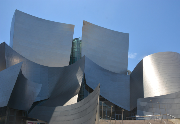 Moderner Konzertsaal mit Glaswänden und Treppen mit Geländern, die zum Eingang führen, umgeben von Laternenmasten, einer Texttafel im Vordergrund und einem bewölkten Himmel.