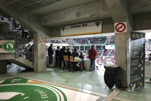 Gruppe von Menschen, die vor einer sitzenden Menge in einem Stadion mit Schildern stehen, die "Boston Celtics vs New York Jets" lesen, ein Müllcontainer, eine Tafel mit Text, eine Treppe mit Geländern und ein Dach mit Deckenleuchten.