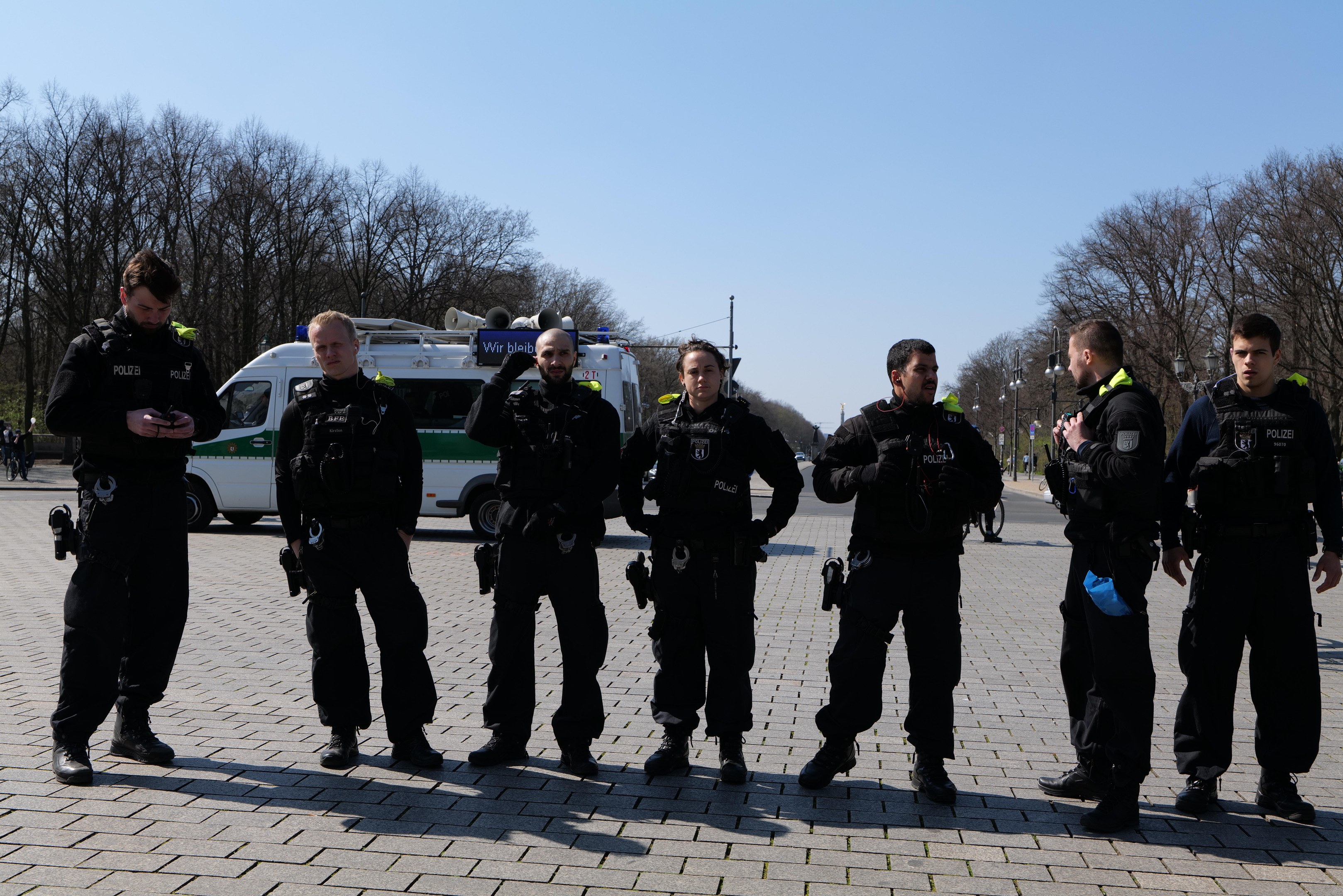 Eine Gruppe von Polizeibeamten steht vor einem Rettungswagen auf einer von Bäumen gesäumten Straße unter einem klaren Himmel.