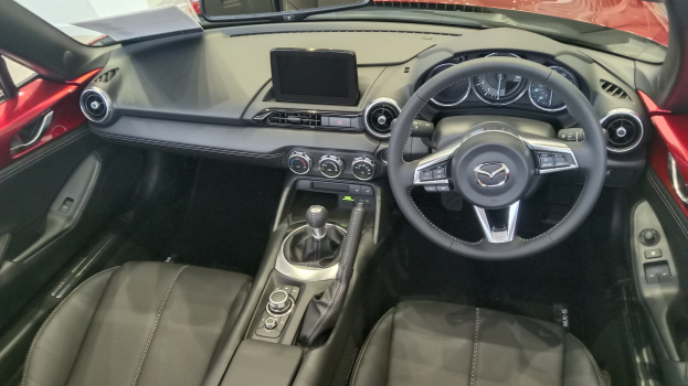Innenansicht eines roten Mazda MX-5 Miata RF mit schwarzen Ledersitzen, Lenkrad, Schaltstange, Knöpfen, Bildschirm, Spiegel und anderen Innenkomponenten.