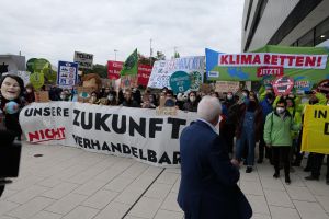 Eine Gruppe von Menschen hält Schilder und Transparente und protestiert gegen den Klimawandel vor einem Gebäude in Deutschland, mit einer Person, die eine Kamera auf der linken Seite hält, Bäumen, Pfählen und einem klaren blauen Himmel im Hintergrund.
