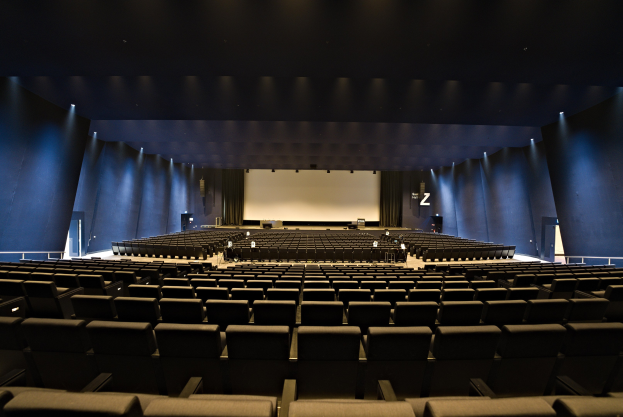 Ein großer Zuschauerraum mit Sitzreihen, wandmontierten Lampen, einer Projektionsleinwand und einigen Gegenständen auf der Bühne.