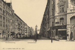 Ein Schwarz-Weiß-Foto einer Stadtstraße in Charlottenburg, Deutschland, mit Gebäuden, Straßenlaternen, Schildern, Kraftfahrzeugen, Fußgängern, Gittern und einem klaren Himmel.