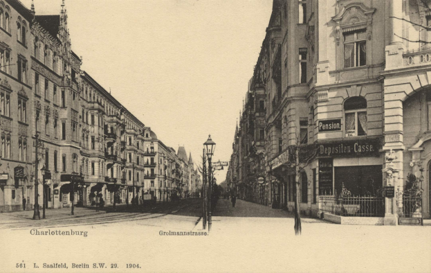 Ein Schwarz-Weiß-Foto einer Stadtstraße in Charlottenburg, Deutschland, mit Gebäuden, Straßenlaternen, Schildern, Kraftfahrzeugen, Fußgängern, Gittern und einem klaren Himmel.