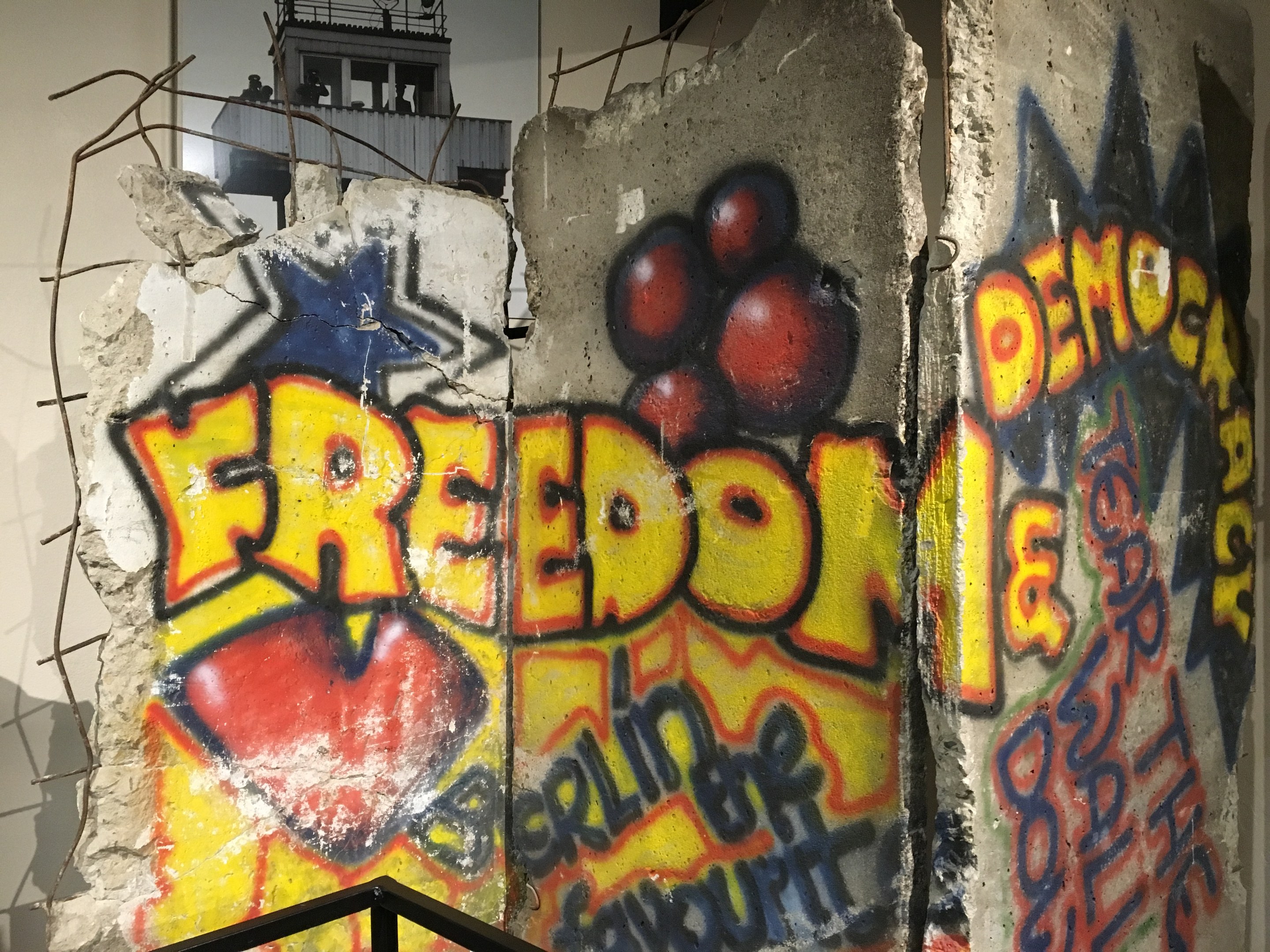 Mauer mit Graffiti, das in fetten schwarzen Buchstaben "Freiheit und Demokratie" liest, ein Geländer unten und ein Gebäude mit Fenstern im Hintergrund.