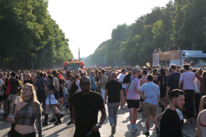 Eine große Menschenmenge, die eine von Bäumen gesäumte Straße entlanggeht, mit einem Turm im Hintergrund und Fahrzeugen mit Menschen auf der rechten Seite, wahrscheinlich beim Christopher Street Day in Berlin.