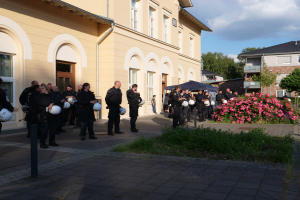 Eine Gruppe von Polizisten steht vor einem Gebäude mit Fenstern und Türen, hält Helme in den Händen und hat in der Nähe ein Zelt aufgebaut, umgeben von Pflanzen mit Blumen, Gras, Bäumen und einem Laternenpfahl unter einem bewölkten Himmel.