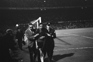 Schwarzes Bild von Fußballspielern, die auf einem Feld mit einem Torpfosten und einer beleuchteten Stadionkulisse im Hintergrund feiern.