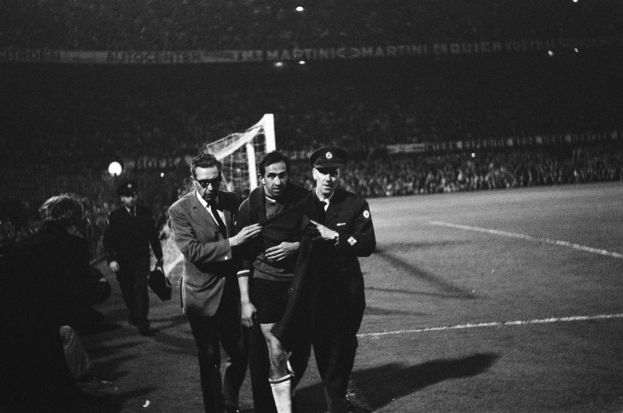 Schwarzes Bild von Fußballspielern, die auf einem Feld mit einem Torpfosten und einer beleuchteten Stadionkulisse im Hintergrund feiern.