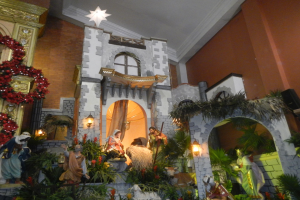 Eine Krippe in einer Kirche, die für Weihnachten geschmückt ist und Statuen von Menschen, Tieren, Pflanzen, Lichtern und anderen Gegenständen im Vordergrund sowie eine Wand und einen Stern im Hintergrund zeigt.