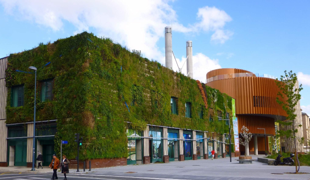 Großes Gebäude mit grüner Wand an der Seite, umgeben von Straßenlaternen, Verkehrspfosten, Verkehrssignalen, Kraftfahrzeugen auf der Straße, Fußgängern auf dem Gehweg, Bäumen und einem Himmel mit Wolken im Hintergrund.