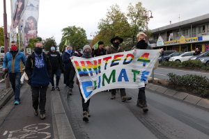 Eine Gruppe von Menschen mit Masken geht eine Straße entlang und hält ein 'United 4 Climate'-Schild hoch, mit einem Gehweg, Geländer, Schildern, Bäumen, Laternen, Fahrzeugen, Gebäuden und einem klaren blauen Himmel im Hintergrund.