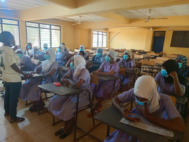 Ein Klassenzimmer in Nigeria mit Schülern, die Gesichtsmasken tragen, auf Bänken sitzen und stehen, mit Büchern und Gegenständen auf Tischen, Fenstern, einer Tür und Deckenleuchten und Ventilatoren im Hintergrund.