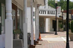Ein Rathaus umgeben von Geb├Ąuden, Laternenmasten, Laternen, Schilden, Kraftfahrzeugen, Absperrgittern, St├╝hlen, Topfpflanzen, B├Ąumen und einem bew├Âlktem Himmel.