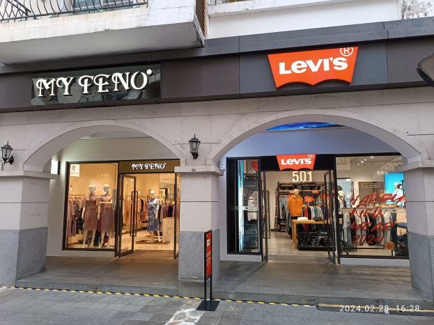 Ladenfront mit einem "Levi's"-Schild, Schaufensterpuppen in Kleidern, hängende Kleidung, Beleuchtung, Texttafeln, Geländer und eine Straße unten.