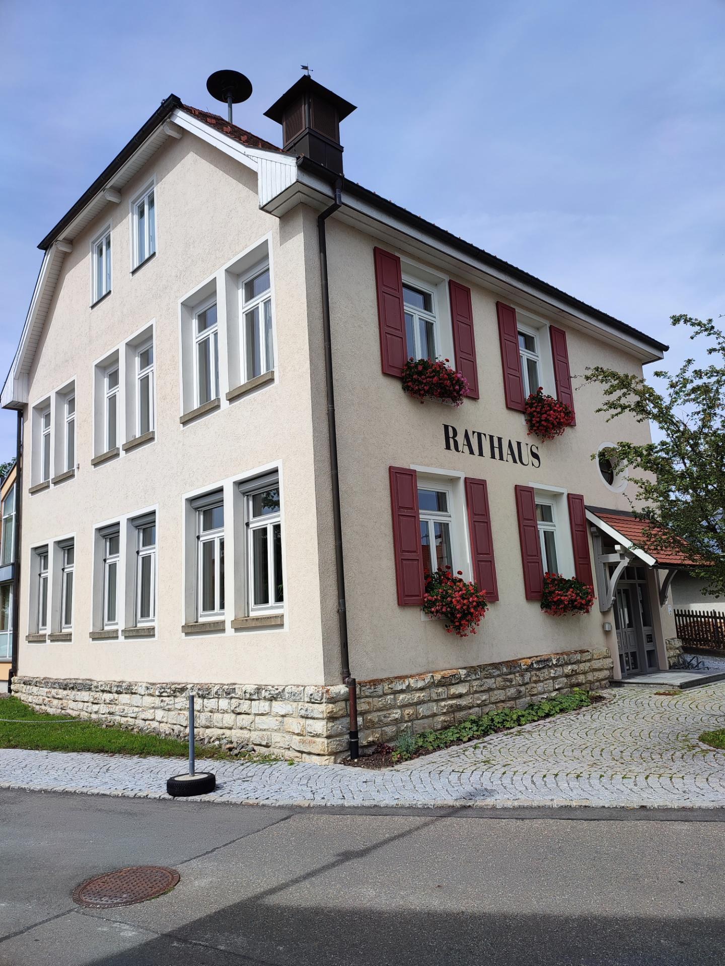 Weißes Gebäude mit roten Fensterläden, umgeben von Grün und Blumen, mit einer Straße unten und Himmel im Hintergrund, identifiziert als das Rathaus-Hotel in Baden-Württemberg.